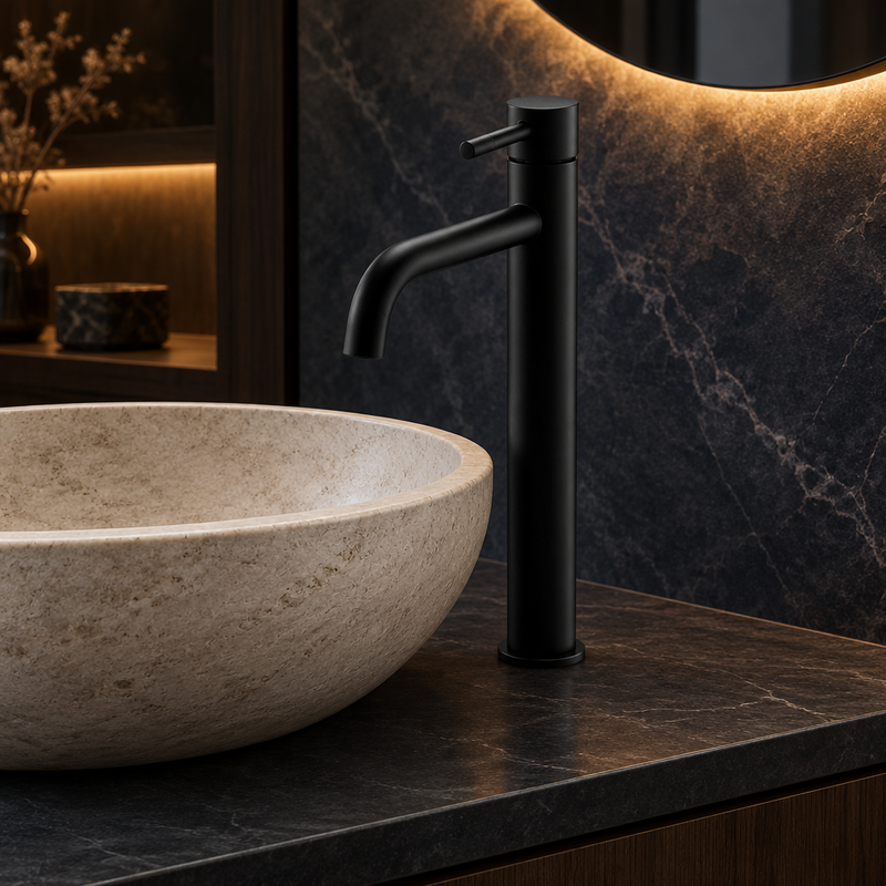 Tall matt-black basin mixer with curved spout installed beside a pale stone vessel basin on a dark countertop in a warm-lit bathroom